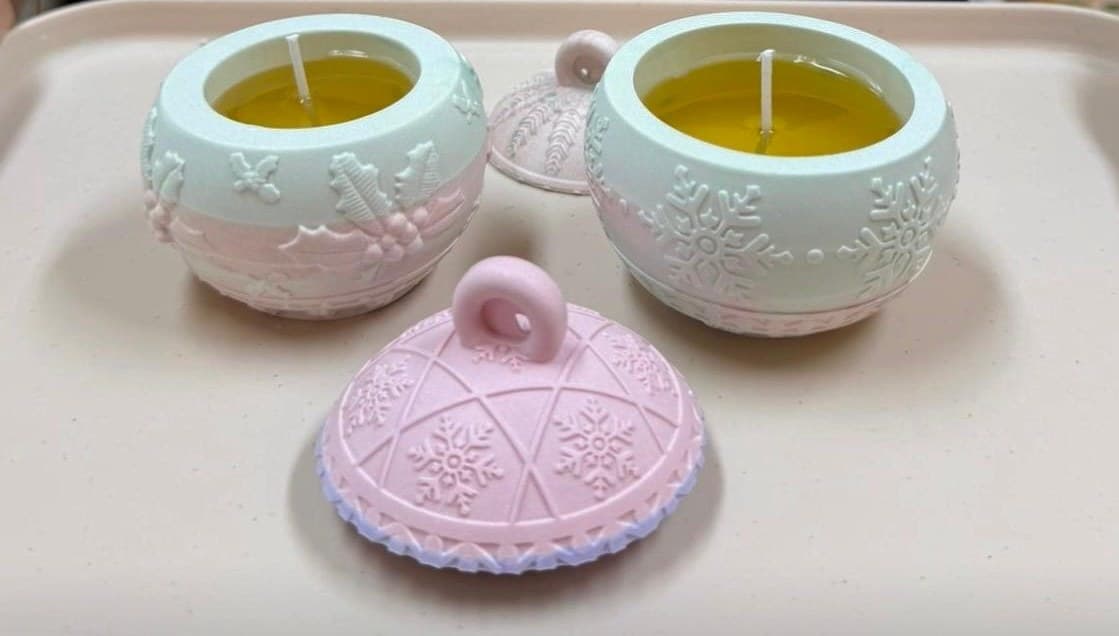 Two pastel Christmas bauble candle jars with snowflake and holly patterns and their lids